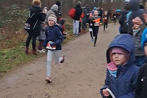 Gruppe von Kindern beim Laufen auf einem Waldweg, Zuschauer applaudieren am Rand. Einige Kinder tragen Startnummern.