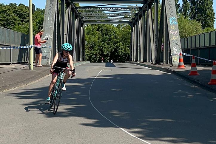 Ein Kind fährt mit einem Fahrrad über eine Stahlbrücke, während ein Erwachsener im Hintergrund steht.