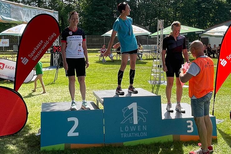 Drei Sportlerinnen stehen auf dem Siegertreppchen des L&ouml;wen Triathlons, w&auml;hrend ein Veranstalter Preisunterlagen &uuml;berreicht.