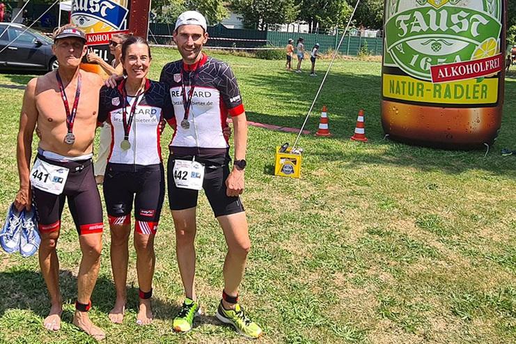 Drei Sportler mit Medaillen posieren vor einem gro&szlig;en, aufblasbaren Fahrradtrinkstand in einem Park.