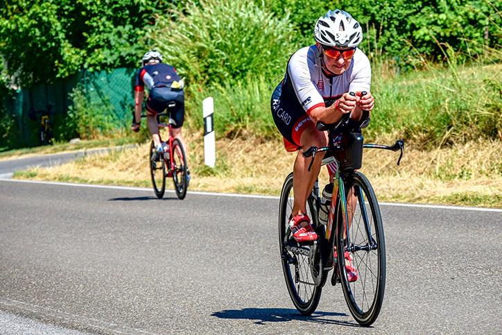 Sportler auf Rennrad fährt auf Straße, während ein weiterer Radfahrer im Hintergrund sichtbar ist. Grüne Landschaft im Hintergrund.