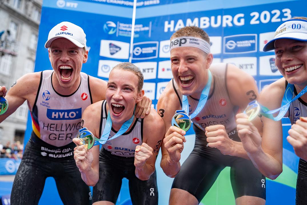 Vier Athleten halten stolz ihre Medaillen vor einem Werbebanner bei der Triathlon-Weltmeisterschaft in Hamburg 2023.
