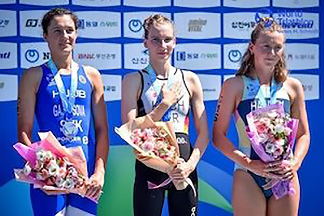 Drei junge Frauen stehen auf dem Siegerpodest, jede mit Medaille und Blumenstrauß vor einer blauen Wand.