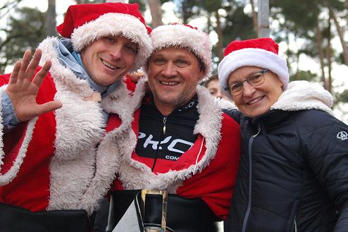 Drei Personen in Weihnachtsmann-Kostümen lächeln und posieren im Freien vor einem Stand und Bäumen.