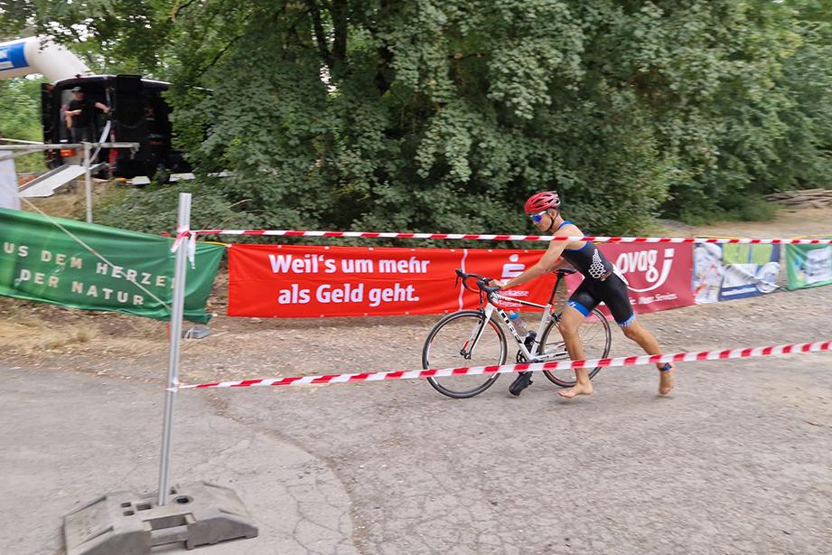 Athlet in einem schwarzen Trisuit schiebt sein Rad vorbei an Werbebannern und einer festlichen Kulisse mit Bäumen.