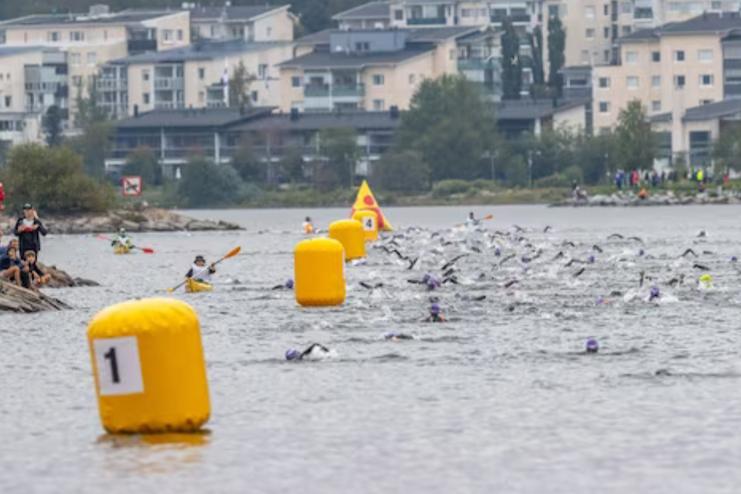 Schwimmer in einem Gew&auml;sser mit gelben Bojen, umgeben von Zuschauern und Wohngeb&auml;uden im Hintergrund.