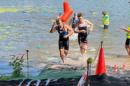 Zwei Athleten laufen aus einem Gew&auml;sser, w&auml;hrend eine Person im Hintergrund sie anfeuert. Sichtbare Absperrungen und Markierungen.
