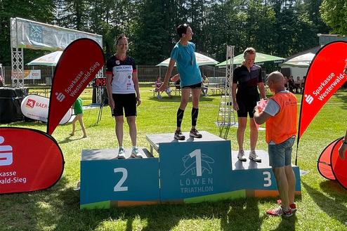 Drei Sportlerinnen stehen auf dem Siegertreppchen des Löwen Triathlons, während ein Veranstalter Preisunterlagen überreicht.