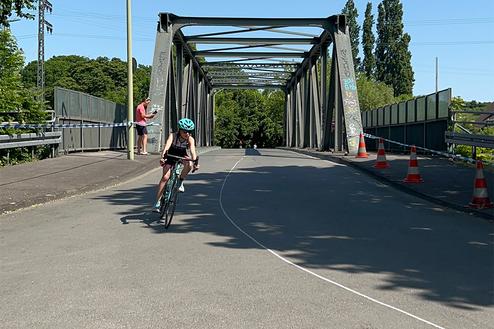 Ein Kind fährt mit einem Fahrrad über eine Stahlbrücke, während ein Erwachsener im Hintergrund steht.