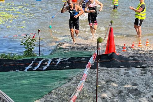 Zwei Athleten laufen aus einem Gewässer, während eine Person im Hintergrund sie anfeuert. Sichtbare Absperrungen und Markierungen.
