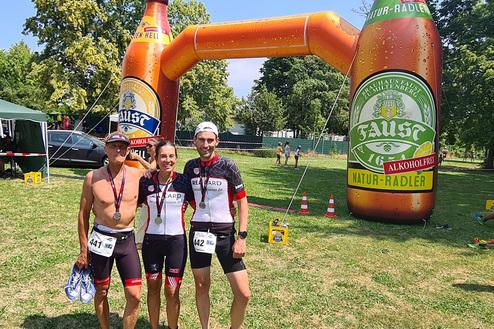 Drei Sportler mit Medaillen posieren vor einem großen, aufblasbaren Fahrradtrinkstand in einem Park.