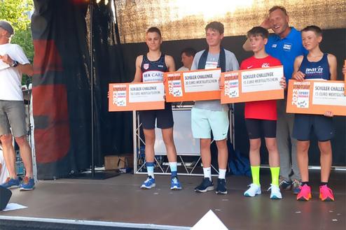 Vier Gewinner der Schüler-Challenge halten ihre Gutscheine auf einer Bühne, während ein Moderator anspricht.