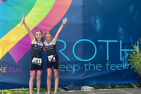 Zwei fröhliche Mädchen in Sportanzügen posieren mit erhobenen Armen vor einer bunten Werbefläche.