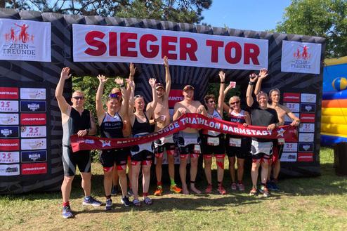 Gruppe von Triathleten feiert unter einem Siegerbogen mit einem Banner für den "Darmstadt Triathlon".