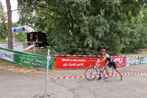 Athlet in einem schwarzen Trisuit schiebt sein Rad vorbei an Werbebannern und einer festlichen Kulisse mit Bäumen.