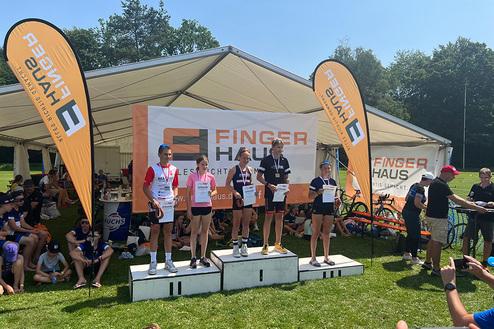 Siegerpodium mit fünf Athleten, Medaillen, Urkunden und Banner in einem festlichen Zelt bei sonnigem Wetter.