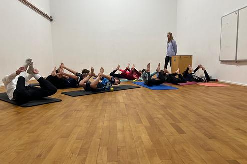Gruppe von Personen auf Matten, die im Fitnessraum Dehnübungen mit angezogenen Füßen ausführen. Eine Lehrerin beobachtet.