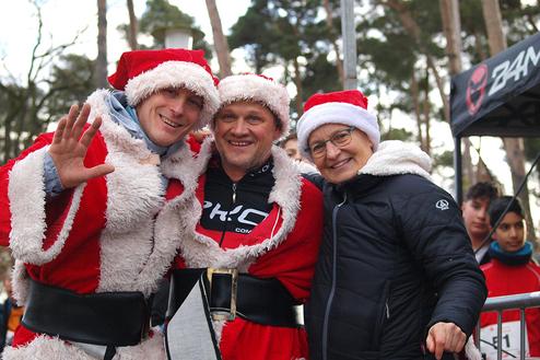 Drei Personen in Weihnachtsmann-Kostümen lächeln und posieren im Freien vor einem Stand und Bäumen.