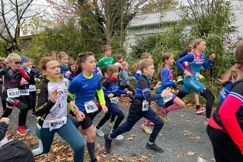 Gruppe von Kindern in Sportkleidung beim Laufen auf einem Weg, umgeben von Bäumen und herbstlichem Laub.