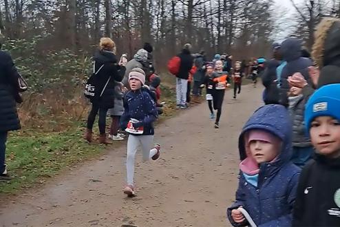 Gruppe von Kindern beim Laufen auf einem Waldweg, Zuschauer applaudieren am Rand. Einige Kinder tragen Startnummern.