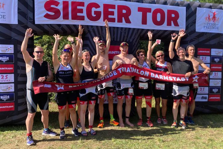 Gruppe von Triathleten feiert unter einem Siegerbogen mit einem Banner f&uuml;r den "Darmstadt Triathlon".
