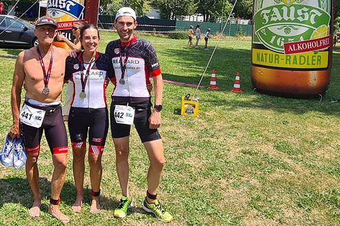 Drei Sportler mit Medaillen posieren vor einem großen, aufblasbaren Fahrradtrinkstand in einem Park.