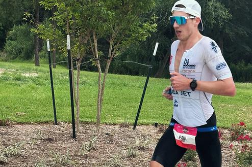 Läufer in sportlicher Kleidung mit Sonnenbrille rennt auf einem gepflasterten Weg, umgeben von Bäumen und Sträuchern.