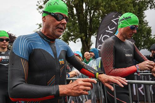 Zwei Triathleten in Neoprenanzügen warten am Start, umgeben von Zuschauern und Fahnenschwenkern bei regnerischem Wetter.