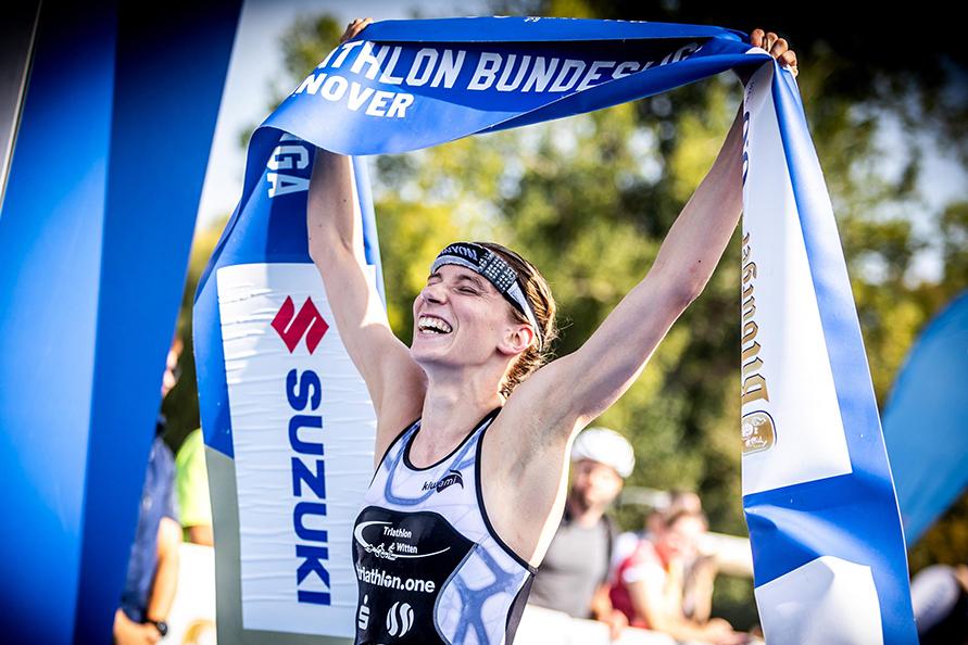 Athlet in Wettkleidung hebt triumphierend das Siegerband beim Triathlon in Hannover über den Kopf.