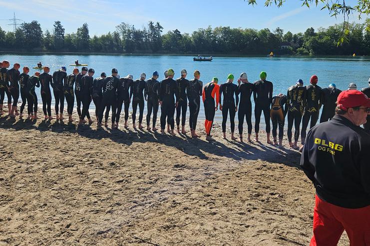 Gruppierung von Schwimmern in Neoprenanz&uuml;gen am Ufer eines Gew&auml;ssers, im Hintergrund Boote und eine sonnige Landschaft.