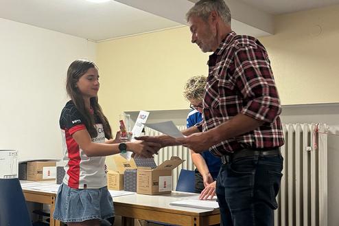 Ein Mädchen überreicht einem Mann ein Dokument bei einer Preisverleihung, weitere Personen und Kartons im Hintergrund.