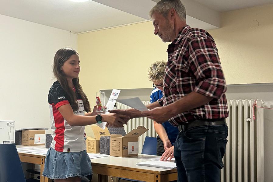 Ein Mädchen überreicht einem Mann ein Dokument bei einer Preisverleihung, weitere Personen und Kartons im Hintergrund.