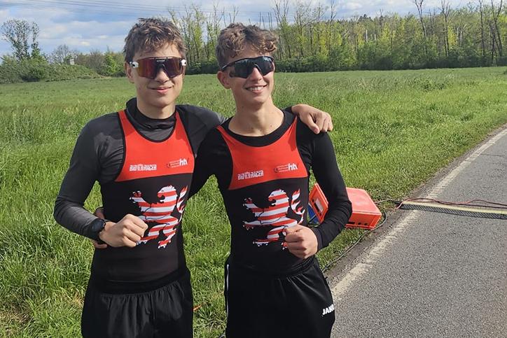 Zwei junge Männer in Sportkleidung stehen lächelnd auf einer Wiese neben einer Landstraße und posieren für ein Foto.