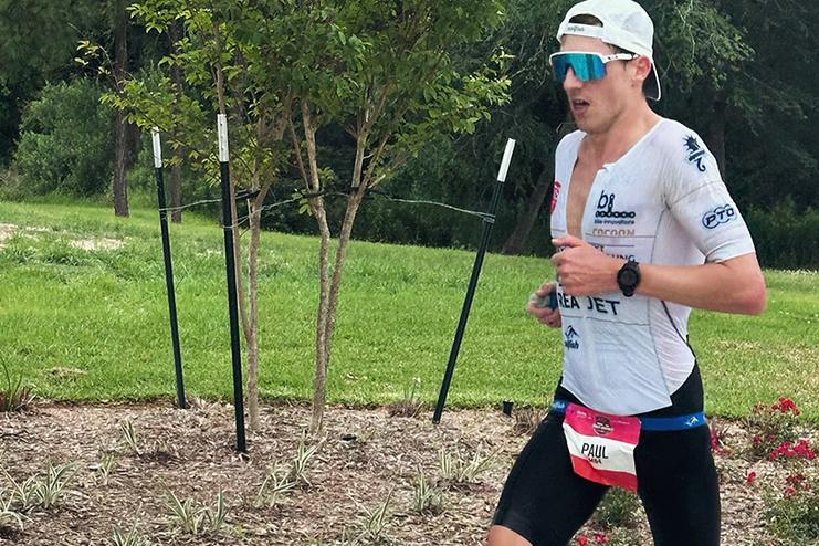 L&auml;ufer in sportlicher Kleidung mit Sonnenbrille rennt auf einem gepflasterten Weg, umgeben von B&auml;umen und Str&auml;uchern.