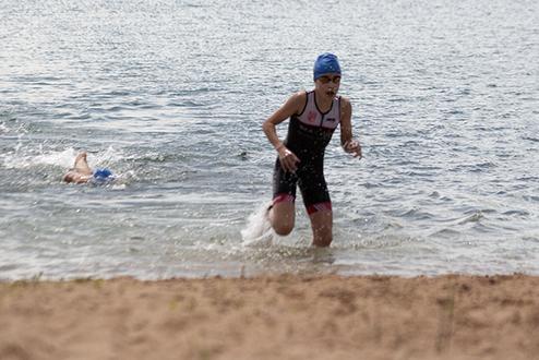 Ein Triathlet in Neoprenanzug läuft aus dem Wasser, während Schwimmer im Hintergrund weiter trainieren.