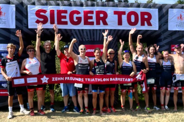 Gruppenfoto von Triathleten mit erhobenen H&auml;nden vor einem Siegerbogen beim &bdquo;Zehn Freunde Triathlon Darmstadt&ldquo;.