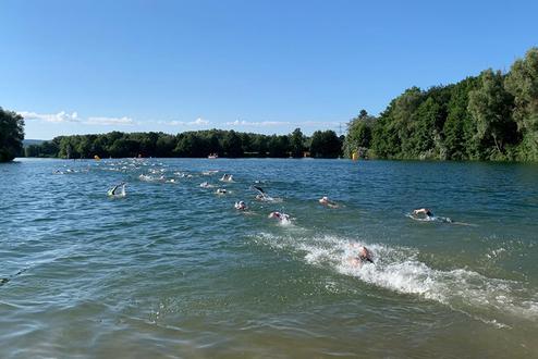 Gruppenschwimmer im klaren Wasser eines Sees, umgeben von Bäumen und blauem Himmel, einige mit Schwimmbojen.