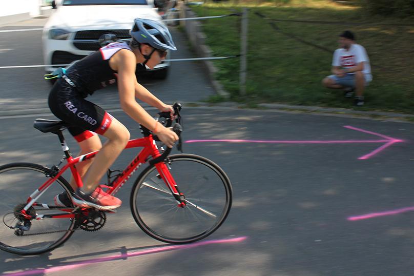 Junge fährt auf einem roten Rennrad, während ein Mann im Hintergrund auf dem Boden sitzt. Pinke Markierungen auf dem Asphalt.
