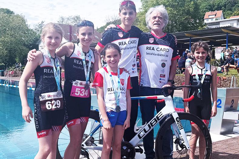Sechs Kinder in Sportbekleidung stehen neben einem Rad vor einem Schwimmbecken bei einem Triathlon-Event.