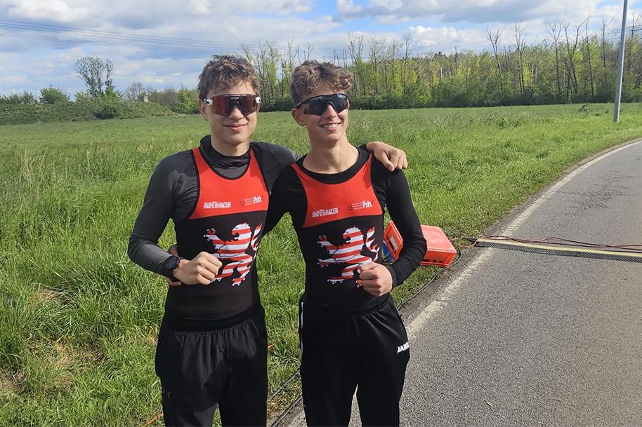Zwei junge Männer in Sportkleidung stehen lächelnd auf einer Wiese neben einer Landstraße und posieren für ein Foto.