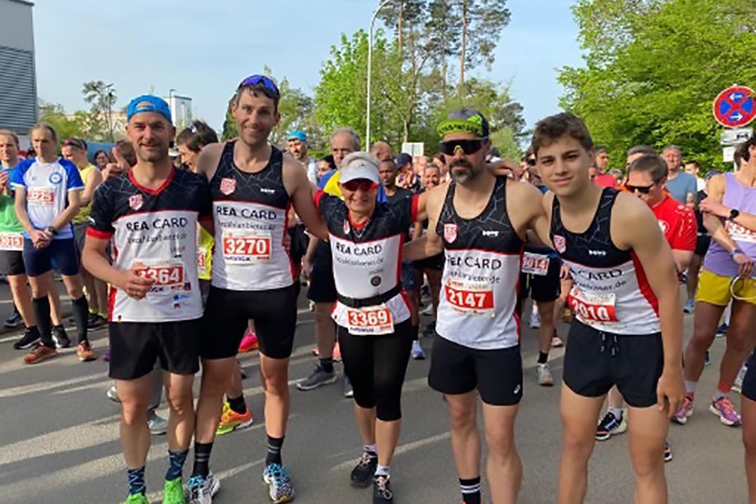 Gruppe von fünf Läufern in Wettkampfkleidung vor dem Start einer Laufveranstaltung mit Zuschauer im Hintergrund.