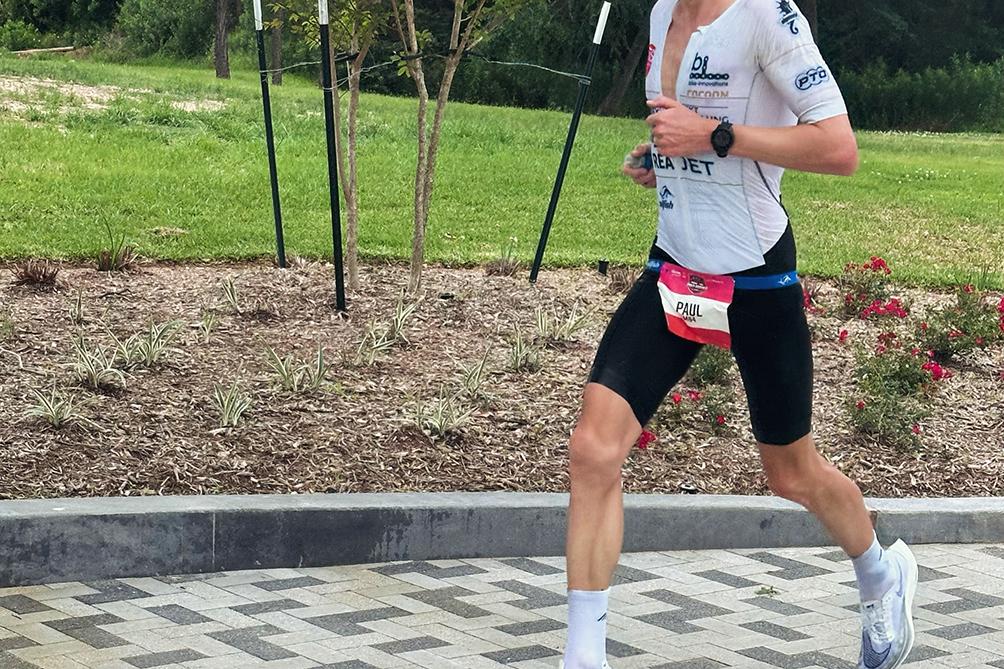 Läufer in sportlicher Kleidung mit Sonnenbrille rennt auf einem gepflasterten Weg, umgeben von Bäumen und Sträuchern.