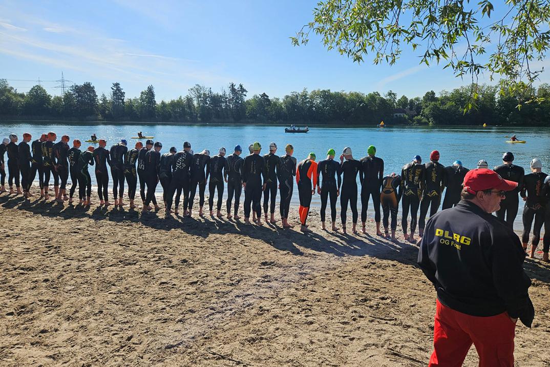 Gruppierung von Schwimmern in Neoprenanzügen am Ufer eines Gewässers, im Hintergrund Boote und eine sonnige Landschaft.