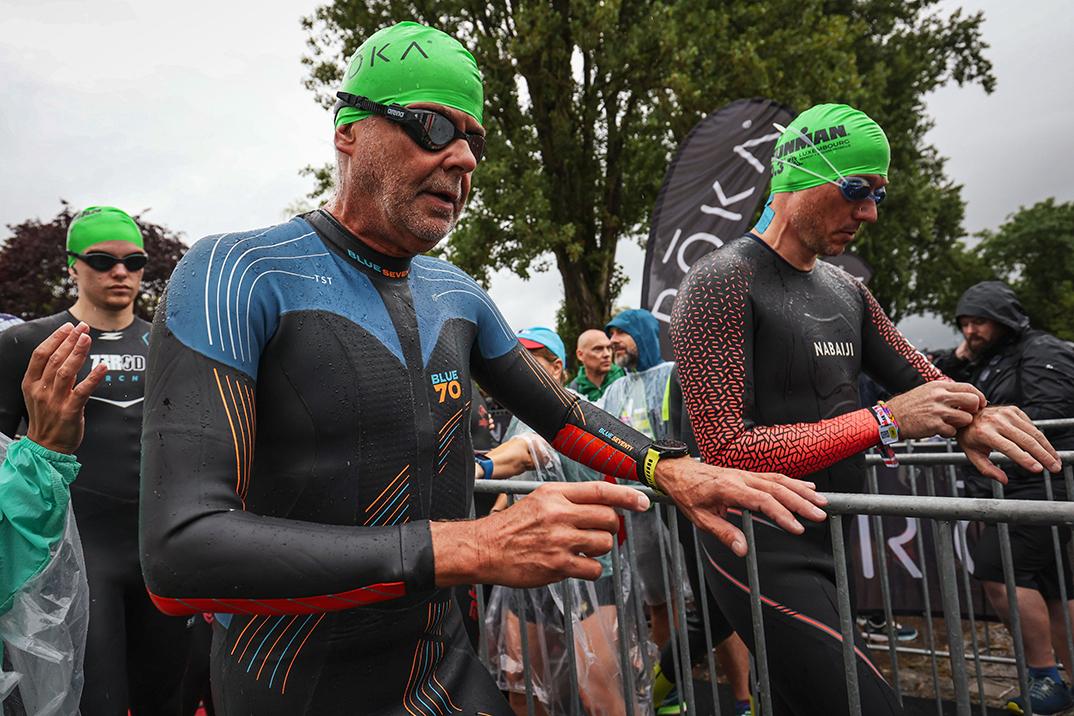 Zwei Triathleten in Neoprenanzügen warten am Start, umgeben von Zuschauern und Fahnenschwenkern bei regnerischem Wetter.