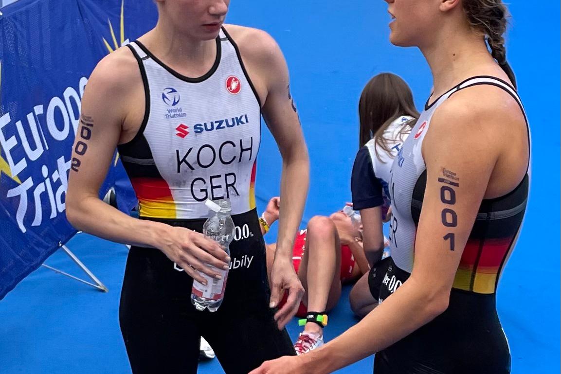 Zwei Triathletinnen im Gespräch, eine hält eine Wasserflasche, beide in sportlicher Bekleidung mit deutschen Nationalfarben.