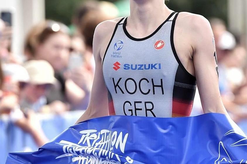 Triathlet überquert die Ziellinie mit blauem Zielband, sichtbar erschöpft, im Hintergrund jubelnde Zuschauer.