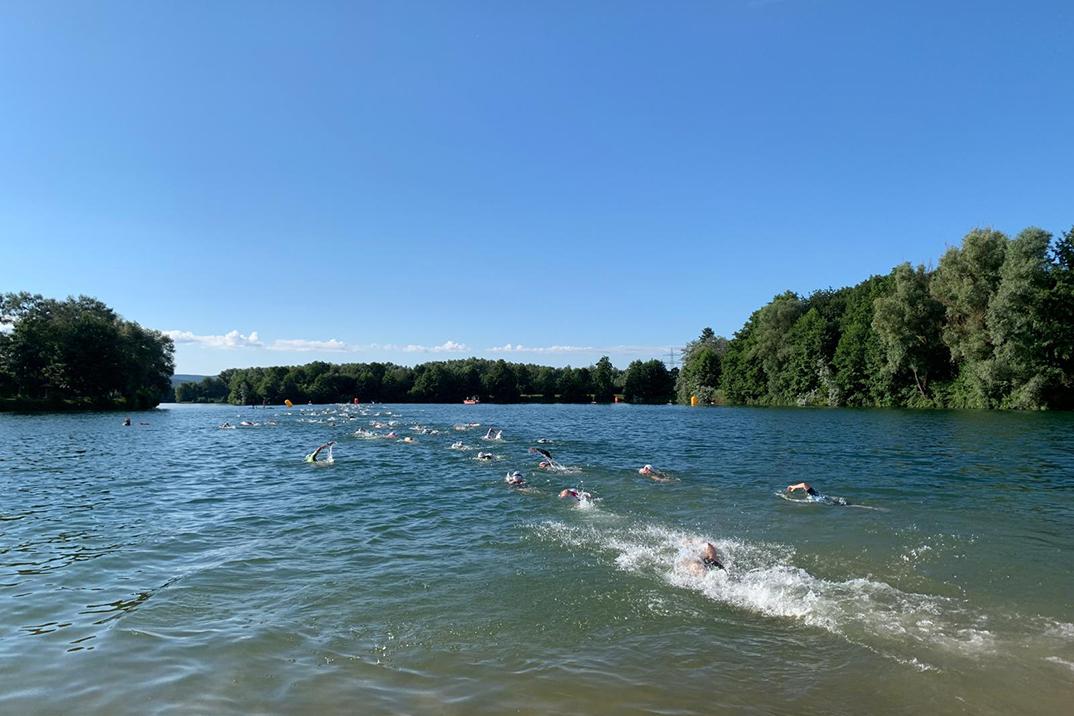 Gruppenschwimmer im klaren Wasser eines Sees, umgeben von Bäumen und blauem Himmel, einige mit Schwimmbojen.