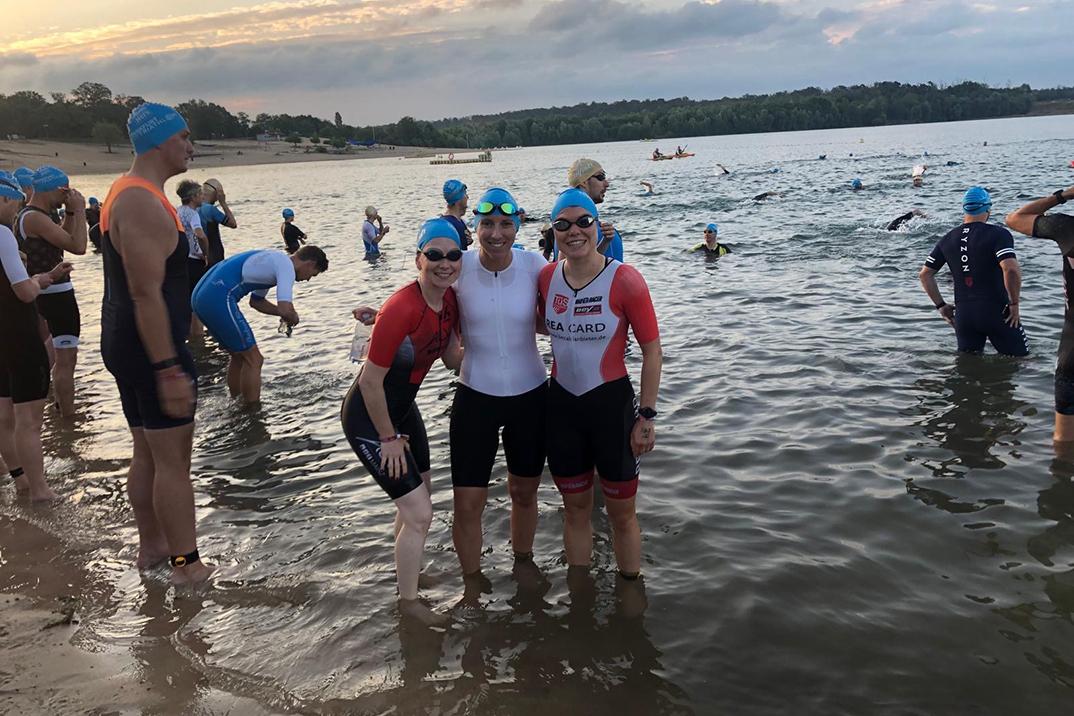 Drei Athletinnen im Wasser lächeln gemeinsam, während im Hintergrund weitere Schwimmer und eine weite Landschaft zu sehen sind.