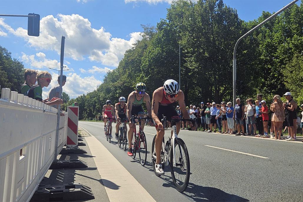 Sechse Triathleten radeln in einem Wettbewerb auf einer Straße, umgeben von Zuschauern unter blauem Himmel.