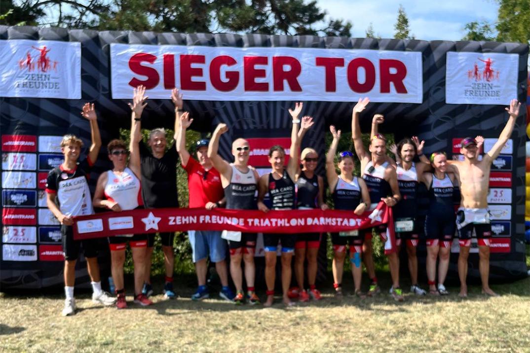 Gruppenfoto von Triathleten mit erhobenen Händen vor einem Siegerbogen beim „Zehn Freunde Triathlon Darmstadt“.